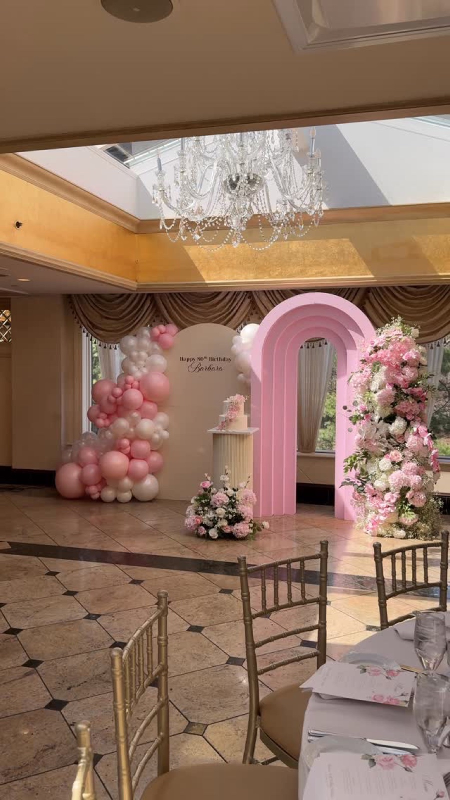 Pink and cream balloon installation by Daydream W.Me at an NJ banquet venue, 80th birthday for Barbara — featuring pink mosaic arch, balloon garland, floral installation, and chandelier in background